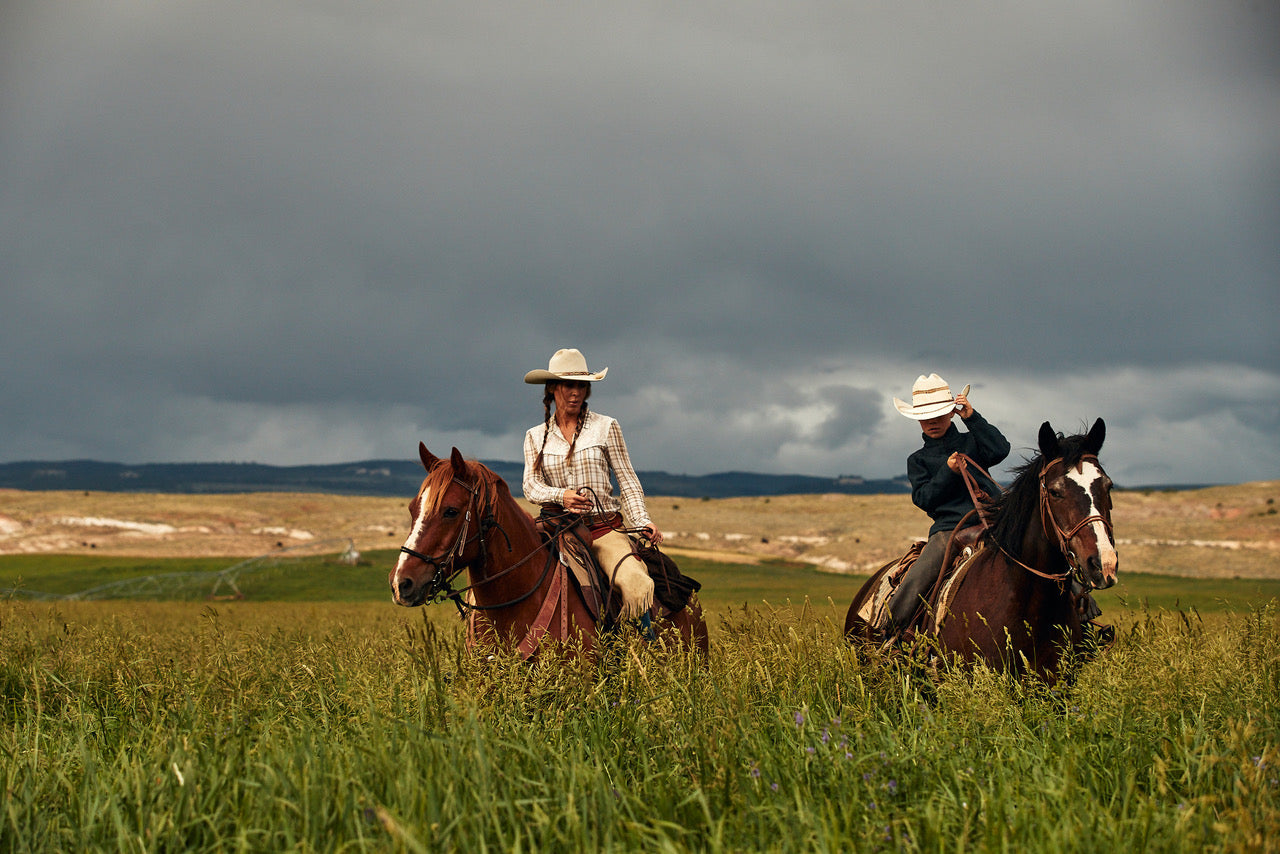 two horseback riders in tall grass with mountains and overcast sky in background