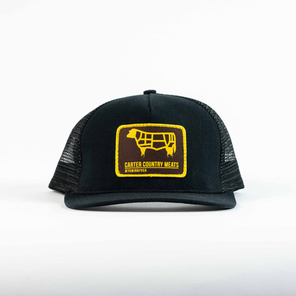 front view black trucker cap with yellow and brown patch that includes cow outline and carter country meats wyoming usa
