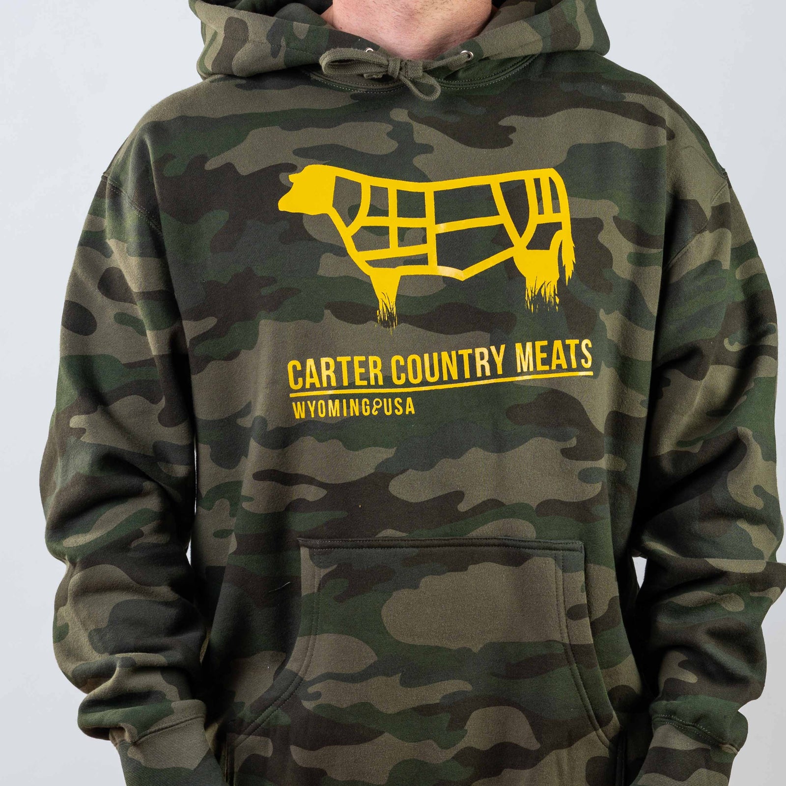 Modeled front view camo hoodie with yellow cow outline and yellow carter country meats wyoming usa text