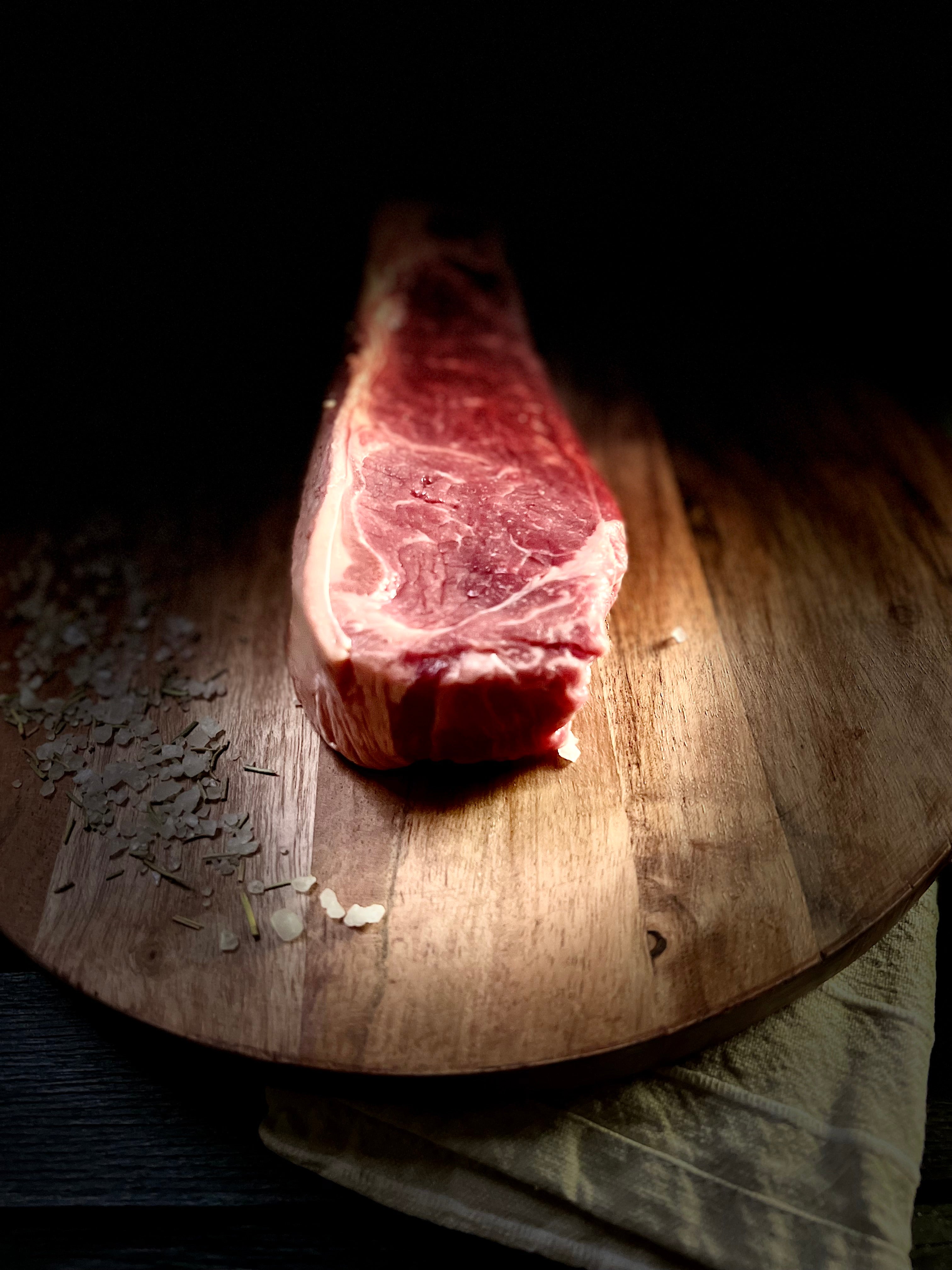 CCM raw NY Strip steak on a round cutting board with herbs